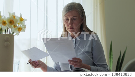 Close up older gray-haired woman sits at a table in the living room, calculating bills and taxes and taking notes. 139413933