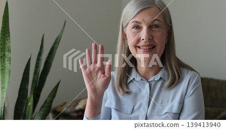 Senior grey-haired woman waving hand talking to webcam video conference calling, online chat meeting at home. Senior grey-haired woman waving hand talking to webcam video conference calling, online chat meeting at home. 139413940