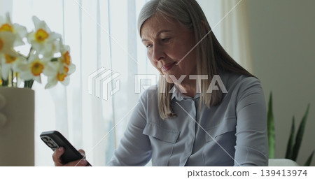 Portrait of senior woman drinking coffee and using smartphone at home. Retirement and senior lifestyle, spending time alone at home. Portrait of senior woman drinking coffee and using smartphone at home. Retirement and senior lifestyle, spending time alone at home. 139413974