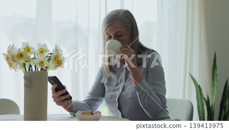 Senior woman drinking coffee and using smartphone for social media, online chat and website at home. 139413975