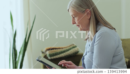 Senior woman browsing digital tablet sitting on sofa in living room at home. Senior woman browsing digital tablet sitting on sofa in living room at home. 139413994
