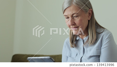 Portrait of serious senior female using TabletPC sitting on sofa at home. Concept of retirement, free time, relax. Portrait of serious senior female using TabletPC sitting on sofa at home. Concept of retirement, free time, relax. 139413996