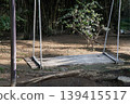 A simple, weathered swing hangs in repose, inviting a moment of rest beneath a verdant canopy where sunlight dances through the garden's leafy shadows. 139415517