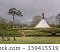 Emerald lawns and sculptural forms rest under the weight of a moody sky, framed by the soft blur of mist-covered mountains. The centerpiece is a striking white pavilion; its tapered crown and conical 139415519