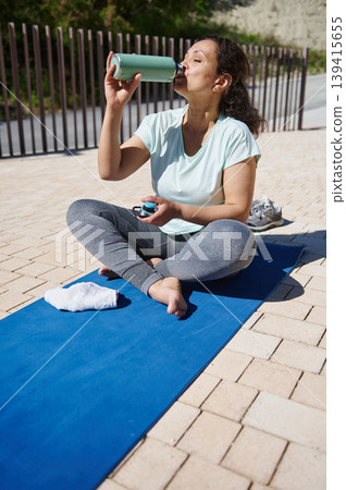 Woman Drinking Water After Outdoor Yoga Session on Blue Mat - Fitness Hydration and Relaxation Woman Drinking Water After Outdoor Yoga Session on Blue Mat - Fitness Hydration and Relaxation 139415655