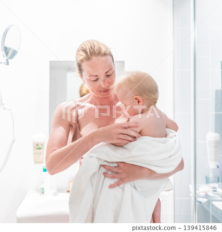 Tender mother holding her baby boy wrapped in a white towel after a bath. Loving connection between a mother and her child in a peaceful moment at home. Tender mother holding her baby boy wrapped in a white towel after a bath. Loving connection between a mother and her child in a peaceful moment at home. 139415846