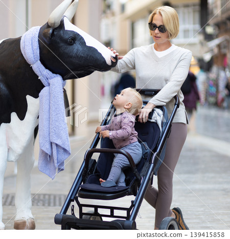 CuriouMother and little son exploring city streets and discovering a funny cow sculptures toddler in a stroller looking at a cow statue in front of a shop in Spain. CuriouMother and little son exploring city streets and discovering a funny cow sculptures toddler in a stroller looking at a cow statue in front of a shop in Spain. 139415858