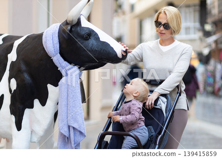CuriouMother and little son exploring city streets and discovering a funny cow sculptures toddler in a stroller looking at a cow statue in front of a shop in Spain. CuriouMother and little son exploring city streets and discovering a funny cow sculptures toddler in a stroller looking at a cow statue in front of a shop in Spain. 139415859