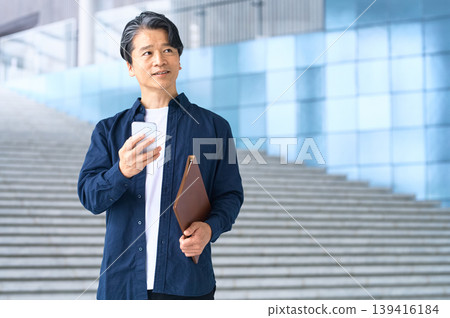 一名五十多歲的日本男子在戶外,拿著智慧型手機,望著遠方。 一名五十多歲的日本男子在戶外,拿著智慧型手機,望著遠方。 139416184