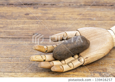 Jointed Wooden Artist Hand Model Holding a Dark wet Rough Stone Used to Exfoliate Skin Lying on a Rustic Weathered Wooden Surface Background Jointed Wooden Artist Hand Model Holding a Dark wet Rough Stone Used to Exfoliate Skin Lying on a Rustic Weathered Wooden Surface Background 139416257