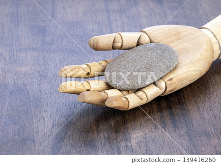Wooden Articulated Artist Hand Model Holding a Smooth Flat Gray River Stone to Exfoliate in a Calming Zen Spa Wellness Concept on Brown Wood Wooden Articulated Artist Hand Model Holding a Smooth Flat Gray River Stone to Exfoliate in a Calming Zen Spa Wellness Concept on Brown Wood 139416260