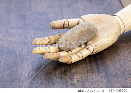 Wooden Artist Mannequin Hand Holding a Smooth River Stone to Exfoliate Skin, Placed on a Rustic Brown Wooden Table Surface for Spa Concept Wooden Artist Mannequin Hand Holding a Smooth River Stone to Exfoliate Skin, Placed on a Rustic Brown Wooden Table Surface for Spa Concept 139416261