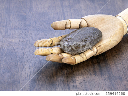 A jointed wooden mannequin hand rests on brown wood surface cradling smooth wet dark basalt stone commonly used to exfoliate and massage skin during hot stone spa treatments A jointed wooden mannequin hand rests on brown wood surface cradling smooth wet dark basalt stone commonly used to exfoliate and massage skin during hot stone spa treatments 139416268