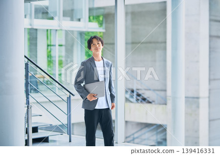 A Japanese man in his 30s stands in the building lobby, holding a laptop computer. A Japanese man in his 30s stands in the building lobby, holding a laptop computer. 139416331