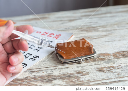 Close-up of a hand with a syringe injecting into a vintage leather wallet against a calendar background, concept of financial injections, healthcare costs, and budget saving. 139416429