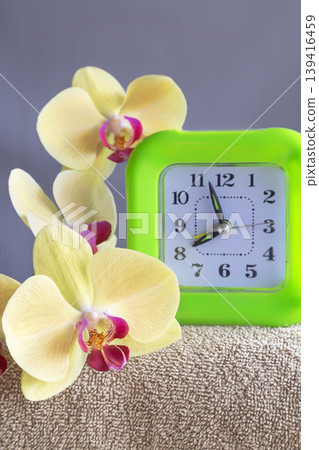 Bright green alarm clock with orchid flowers on soft towel for morning routine concept. Bright green alarm clock with orchid flowers on soft towel for morning routine concept. 139416459