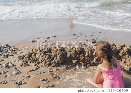 Happy Little Girl in Pink Swimsuit Building Sand Castle Moat on a Pebble Beach with Waves and Sea Foam in Summer Sunlight Happy Little Girl in Pink Swimsuit Building Sand Castle Moat on a Pebble Beach with Waves and Sea Foam in Summer Sunlight 139416709