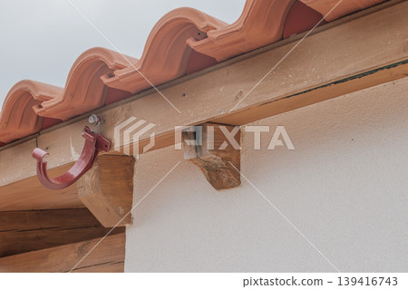 A detailed shot of water damage on a wooden roof beam due to a missing gutter system. The image shows wood rot, darkening, and a lone metal bracket on a tiled roof A detailed shot of water damage on a wooden roof beam due to a missing gutter system. The image shows wood rot, darkening, and a lone metal bracket on a tiled roof 139416743
