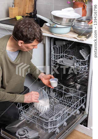 A man is at work in the kitchen, pouring salt into a dishwasher as he prepares to wash dirty dishes. He is focused on the task at hand while organizing the appliances A man is at work in the kitchen, pouring salt into a dishwasher as he prepares to wash dirty dishes. He is focused on the task at hand while organizing the appliances 139416796