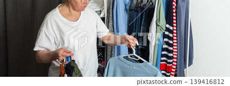 Woman sorts and hangs washed clothes on a hanger. She holds a blue sweatshirt and several other items in her hands. This scene takes place in a home environment, banner 139416812