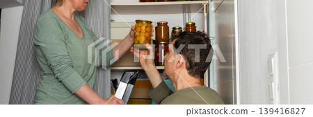 Married couple places jars of canned goods onto shelves in their pantry. They are inside their home with a view of flowers and the city from the balcony. It is autumn, banner Married couple places jars of canned goods onto shelves in their pantry. They are inside their home with a view of flowers and the city from the balcony. It is autumn, banner 139416827