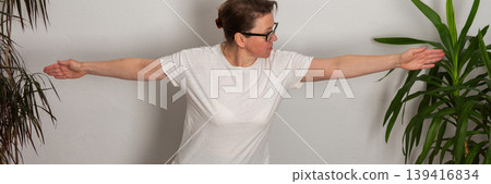A 52-year-old woman is doing yoga at home. She stands on a mat with her arms extended, showing focus and balance. Plants surround her in the room, creating an active environment, banner A 52-year-old woman is doing yoga at home. She stands on a mat with her arms extended, showing focus and balance. Plants surround her in the room, creating an active environment, banner 139416834