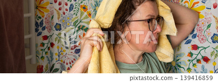 A woman performs her morning bathroom routine. She stands with a towel on her head, looking in the mirror. The colorful floral wall adds brightness to the space, banner A woman performs her morning bathroom routine. She stands with a towel on her head, looking in the mirror. The colorful floral wall adds brightness to the space, banner 139416859