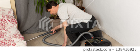 A 53-year-old man uses a vacuum cleaner to clean the floor in a room decorated with houseplants. He focuses on keeping the space tidy and organized while working, banner A 53-year-old man uses a vacuum cleaner to clean the floor in a room decorated with houseplants. He focuses on keeping the space tidy and organized while working, banner 139416923