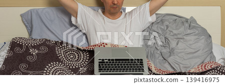 A man is sitting on a bed in a bedroom. He is wrapped in blankets and holding a laptop. He looks frustrated and tired in the early morning light, banner A man is sitting on a bed in a bedroom. He is wrapped in blankets and holding a laptop. He looks frustrated and tired in the early morning light, banner 139416975