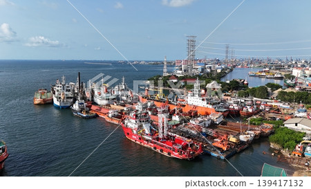 A red offshore supply and rescue ship equipped with a lattice tower, docked at a busy industrial port alongside various barges and heavy maritime equipment. A red offshore supply and rescue ship equipped with a lattice tower, docked at a busy industrial port alongside various barges and heavy maritime equipment. 139417132