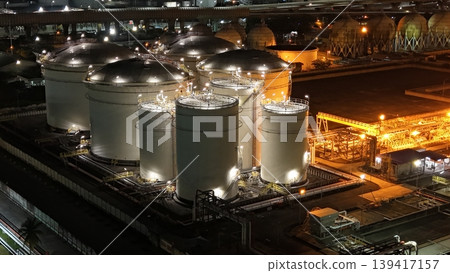 High-angle night view of a fuel storage tank terminal with multiple large, illuminated cylinders and a complex piping infrastructure at an industrial port facility, glowing in the dark. High-angle night view of a fuel storage tank terminal with multiple large, illuminated cylinders and a complex piping infrastructure at an industrial port facility, glowing in the dark. 139417157