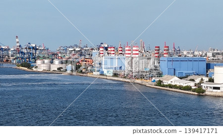 Aerial view of a coastal natural gas power plant station with striped industrial chimneys, high voltage transmission towers, and electrical infrastructure against a clear blue sky. Aerial view of a coastal natural gas power plant station with striped industrial chimneys, high voltage transmission towers, and electrical infrastructure against a clear blue sky. 139417171