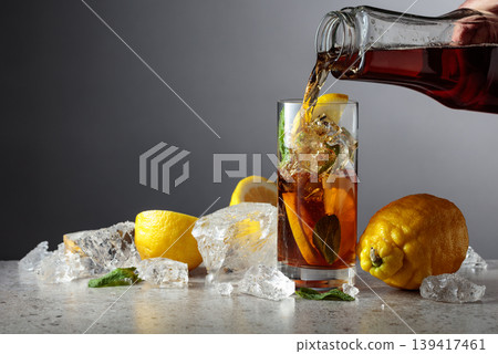 Iced black tea is poured from a bottle into a tall glass with lemon wedges, ice cubes and mint leaves. Iced black tea is poured from a bottle into a tall glass with lemon wedges, ice cubes and mint leaves. 139417461