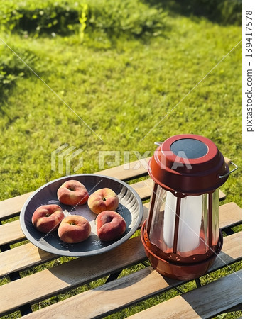 Flat peaches in a metal bowl on a wooden table with a red lantern nearby, creating a seasonal fruit still in a rustic setting 139417578