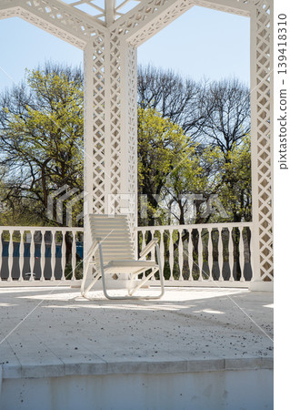 White lounge chair stands on terrace under decorative pavilion with columns and lattice structure overlooking trees. Outdoor furniture, architectural detail, relaxation space, garden design, lifestyle White lounge chair stands on terrace under decorative pavilion with columns and lattice structure overlooking trees. Outdoor furniture, architectural detail, relaxation space, garden design, lifestyle 139418310