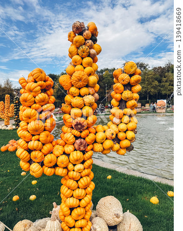 Unique sculpture made of stacked pumpkins resembling a cactus, surrounded by lush greenery and a serene water feature, showcasing autumn creativity and artistry Unique sculpture made of stacked pumpkins resembling a cactus, surrounded by lush greenery and a serene water feature, showcasing autumn creativity and artistry 139418669