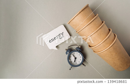 Stack of eco-friendly paper cups beside a vintage alarm clock and a speech bubble with the word energy, symbolizing sustainability and environmental awareness in daily life Stack of eco-friendly paper cups beside a vintage alarm clock and a speech bubble with the word energy, symbolizing sustainability and environmental awareness in daily life 139418681