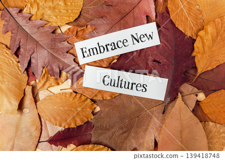 Colorful autumn leaves in various shades of orange, red, and brown surround two white paper strips with the words "Embrace New Cultures" creating a warm seasonal atmosphere Colorful autumn leaves in various shades of orange, red, and brown surround two white paper strips with the words "Embrace New Cultures" creating a warm seasonal atmosphere 139418748