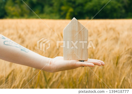 Hand holding wooden house model in a golden wheat field under bright sunlight with greenery Hand holding wooden house model in a golden wheat field under bright sunlight with greenery 139418777