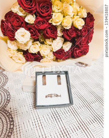 Elegant jewelry set featuring a silver ring and earrings displayed beside a beautiful bouquet of red and white roses, perfect for special occasions and celebrations 139418807