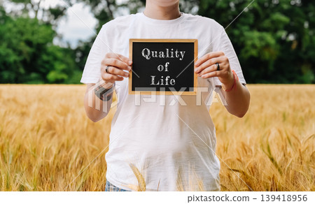 Individual holding chalkboard sign reading "Quality of Life" in golden wheat field with greenery Individual holding chalkboard sign reading "Quality of Life" in golden wheat field with greenery 139418956