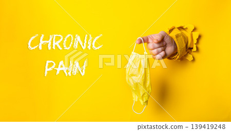 Hand holding yellow mask emerging from torn paper with chronic pain concept on bright background 139419248