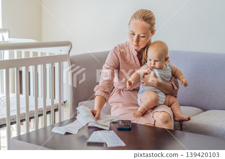 Mother sitting on sofa holding baby while calculating monthly expenses with receipts at home. Stressed mom managing budget and childcare. Financial stress, parenthood and real life concept 139420103