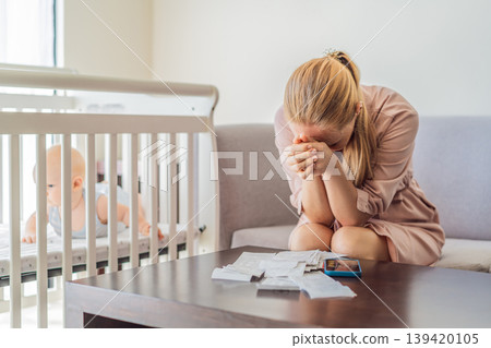 Mother sitting on sofa calculating monthly expenses with receipts while baby lies in crib nearby at home. Stressed mom managing budget and childcare. Financial pressure and parenthood concept 139420105