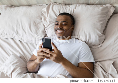 African american man lying in bed using smartphone and smiling African american man lying in bed using smartphone and smiling 139420341