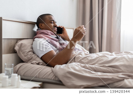 Black man lying in bed talking on phone while checking thermometer Black man lying in bed talking on phone while checking thermometer 139420344