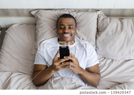 African american man lying in bed smiling while using smartphone African american man lying in bed smiling while using smartphone 139420347