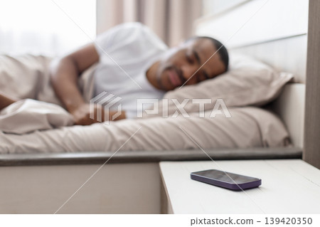 Smartphone on bedside table with black man sleeping in background in bedroom 139420350