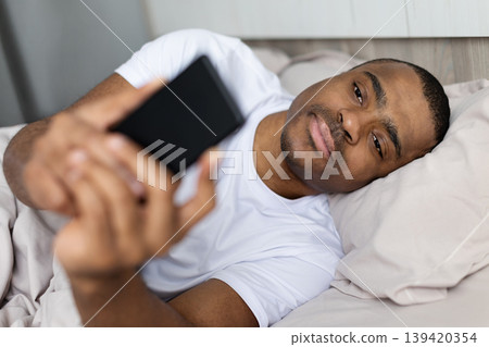 Black man lying in bed using smartphone, looking at screen in bedroom Black man lying in bed using smartphone, looking at screen in bedroom 139420354