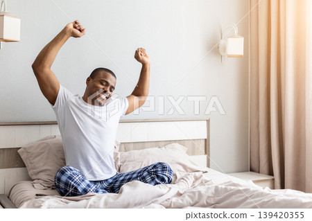 African american man stretching arms while sitting in bed in bedroom 139420355